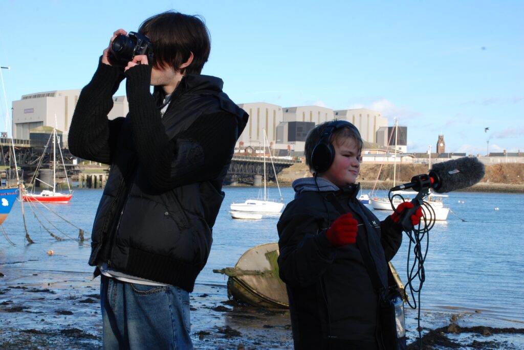 Two teenagers, using cameras, the right is looking into the distance at the right and the left is looking in the distance to the left. In the background the BAE Shipyard is visible and the water in the midground